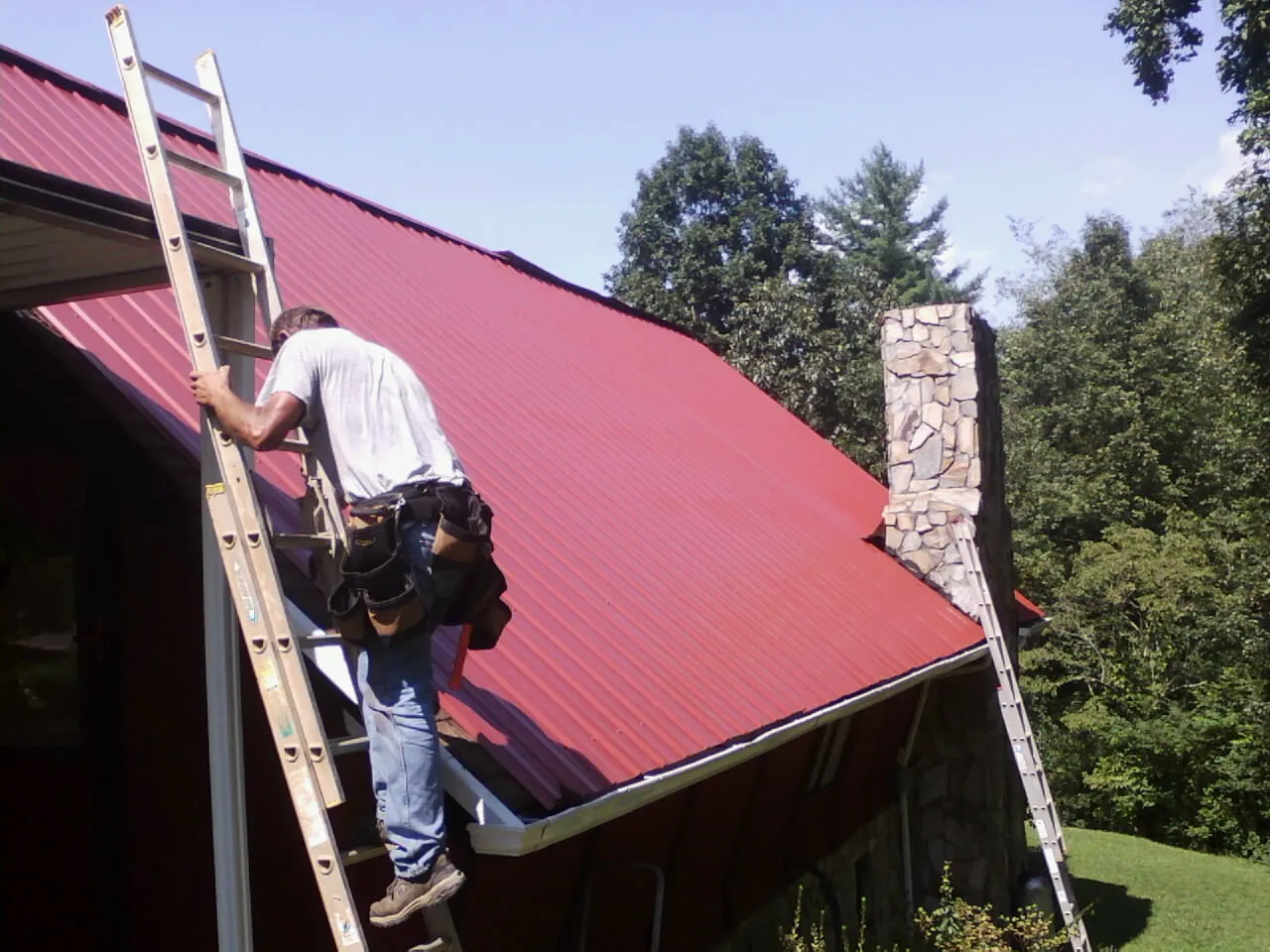 Roofer on a ladder beside a red metal roof and stone chimney — Soffit & Fascia Repair by Roofing Brothers in Gazelle